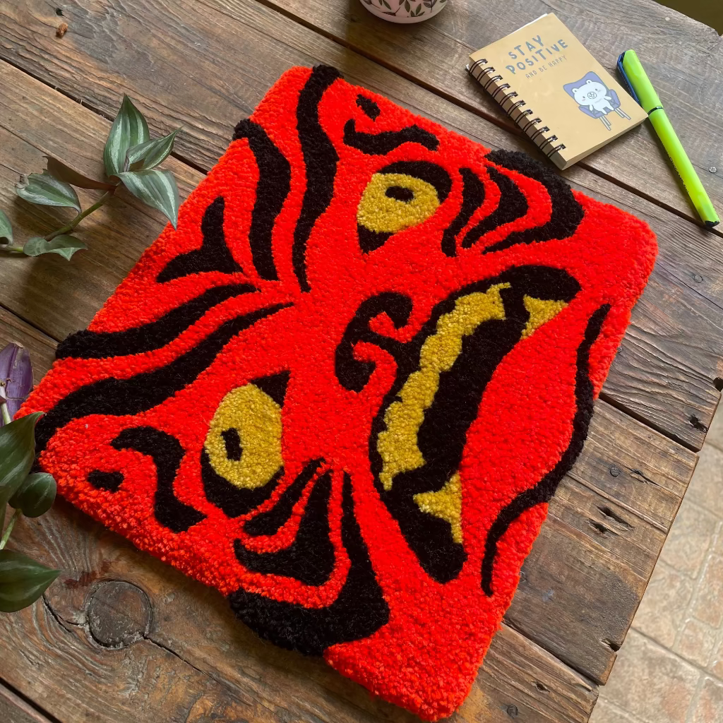 A red and black textured rug with a stylized face design, styled on a rustic wooden table with a mug, notebook, and green pen.